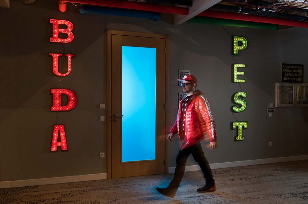A Google Budapest iroda világító Buda és Pest feliratokkal a falakon. A mennyezeten színes csövek futnak, a két felirat által közrezárt üvegbetétes ajtó előtt pedig épp a Google csapatának egyik tagja sétál el.