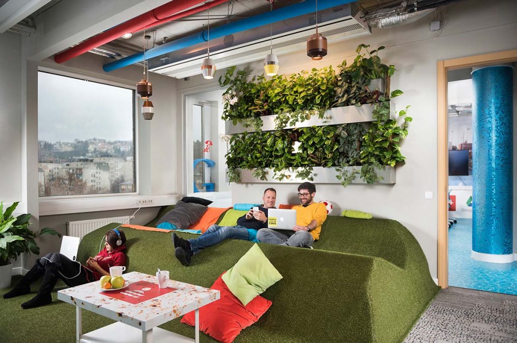 A Google Budapest iroda közösségi tere zöld, füvet imitáló pihenővel, stílusos világítással és növényekkel a falon. Néhány alkalmazott beszélget és relaxál színes párnák között.