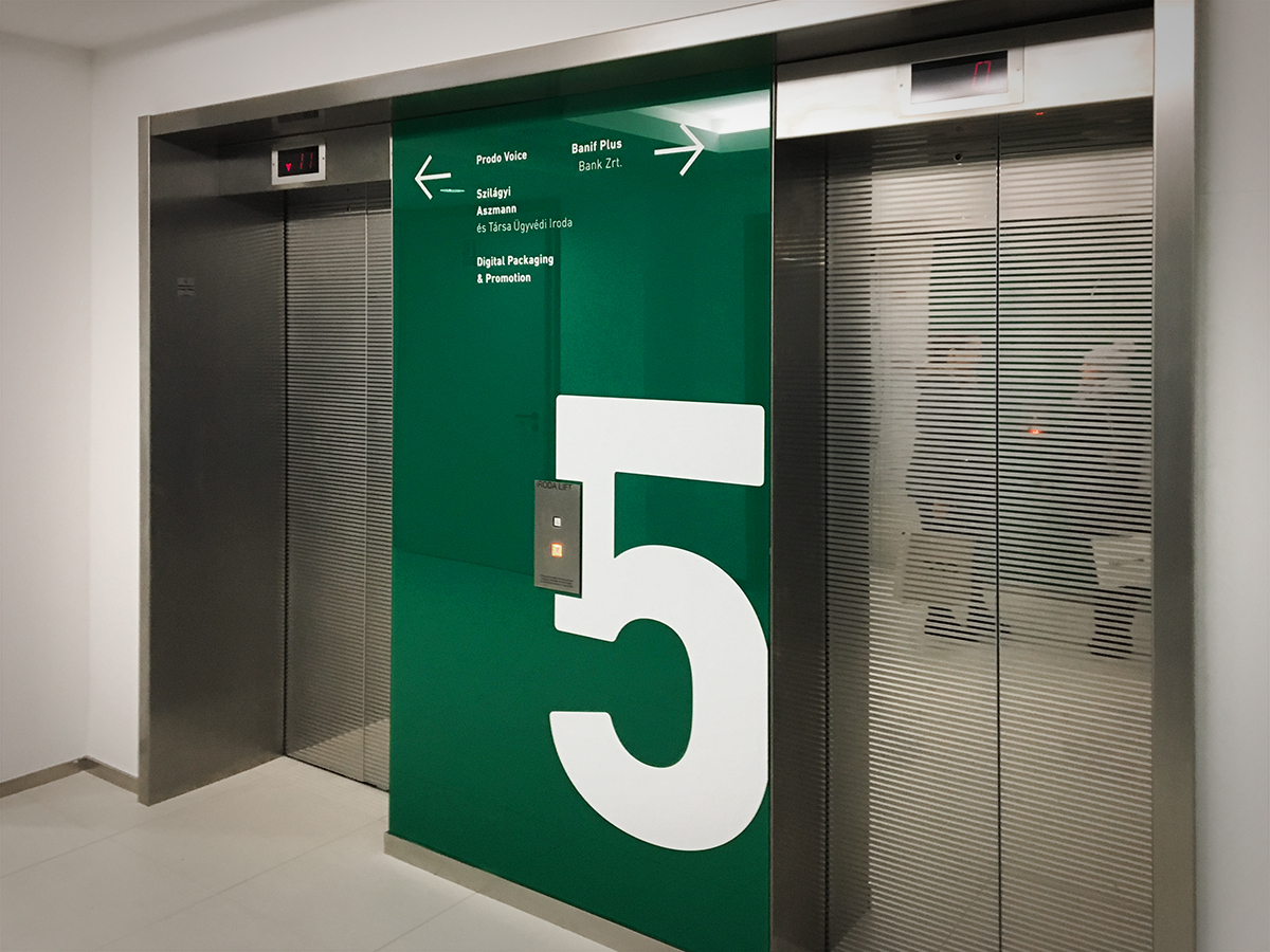 Két liftajtó között lévő nagyméretű, sötétzöld alapon hatalmas fehér 5-ös számmal ellátott irányítótábla. A rajta lévő feliratok segítenek megtalálni az irodákat az adott emeleten.