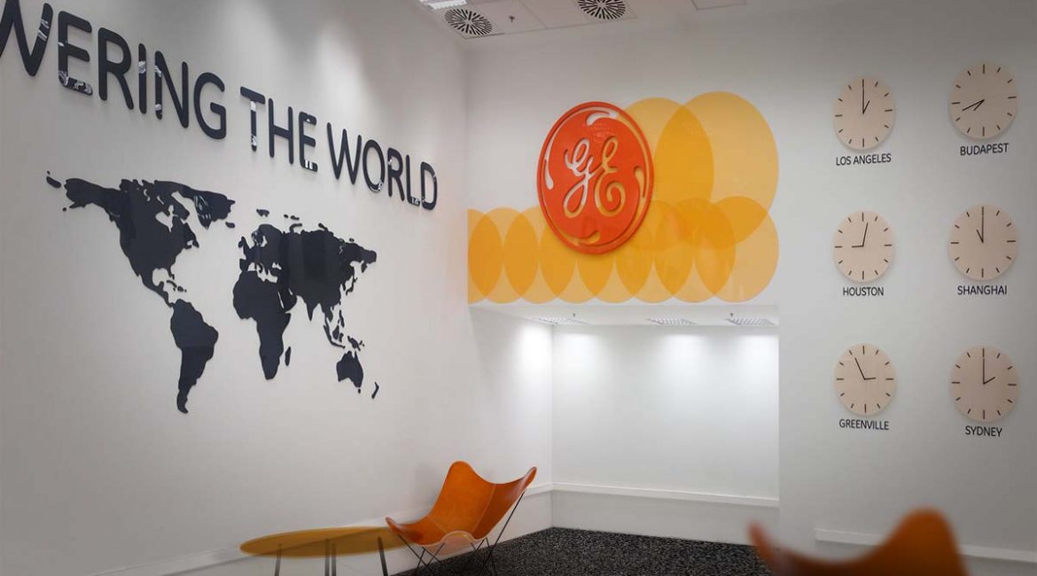 General Electric 7-es épület váróterme nagyméretű "Powering the World" plasztikus felirattal, világtérképpel, a cég nagyméretű logójával és világórákkal a falon.